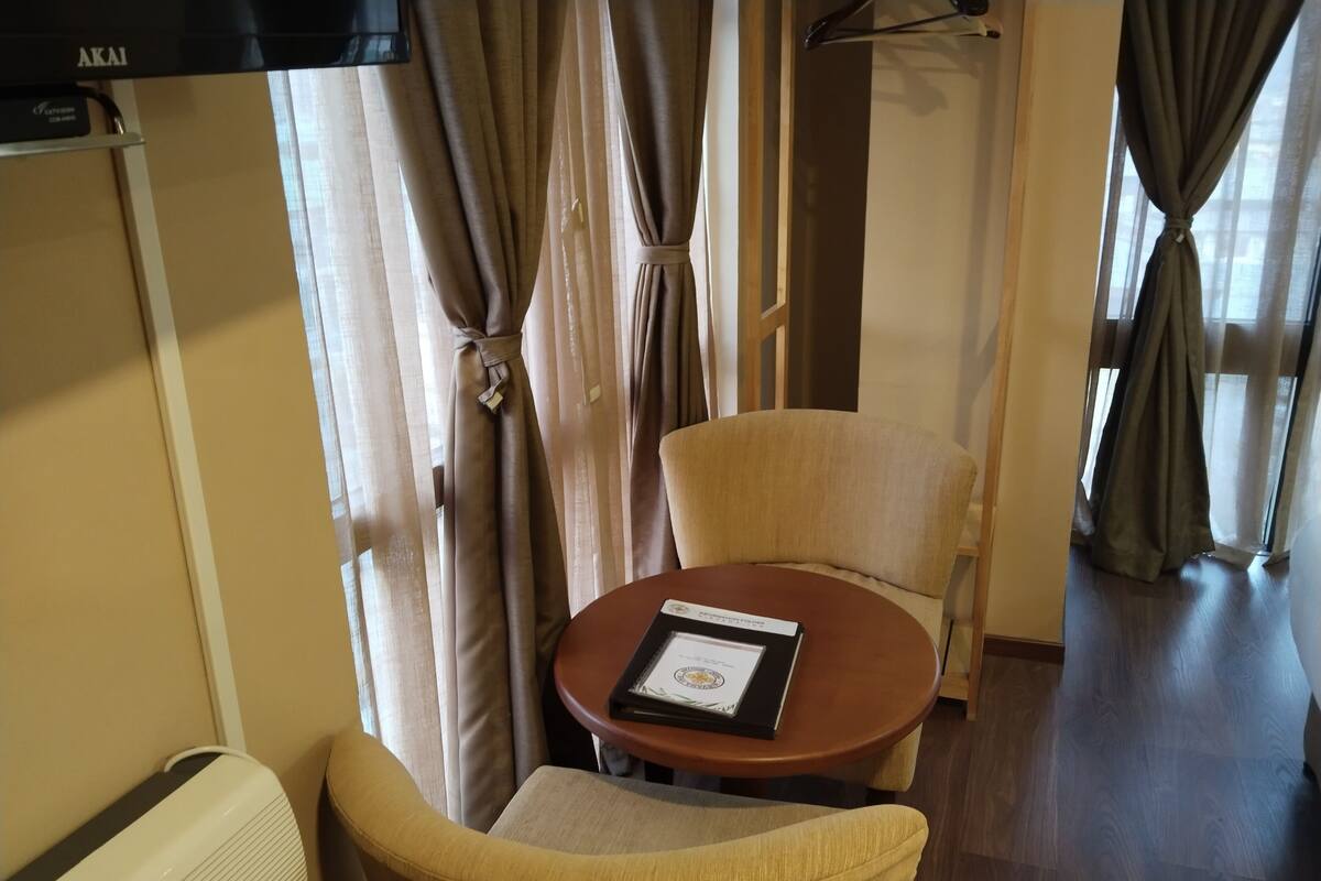 A hotel room with a round table, two armchairs, a wall-mounted heater, and a window with curtains.