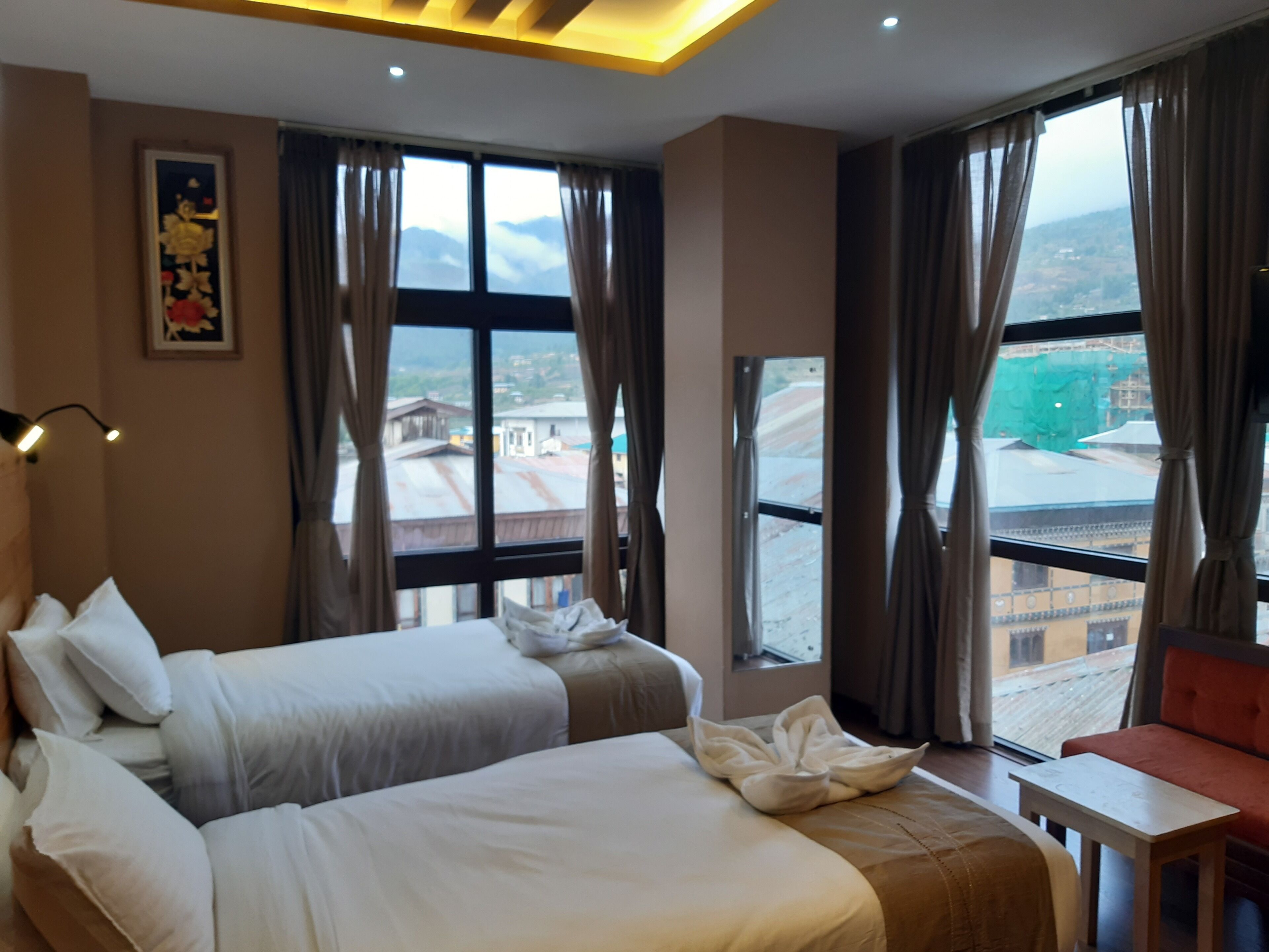 A hotel room with two beds, a small table, and a view of the cityscape through large windows.