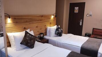 A hotel room with two single beds, wooden headboards, bedside tables with lamps, a door with a lock, and a small table with a chair.