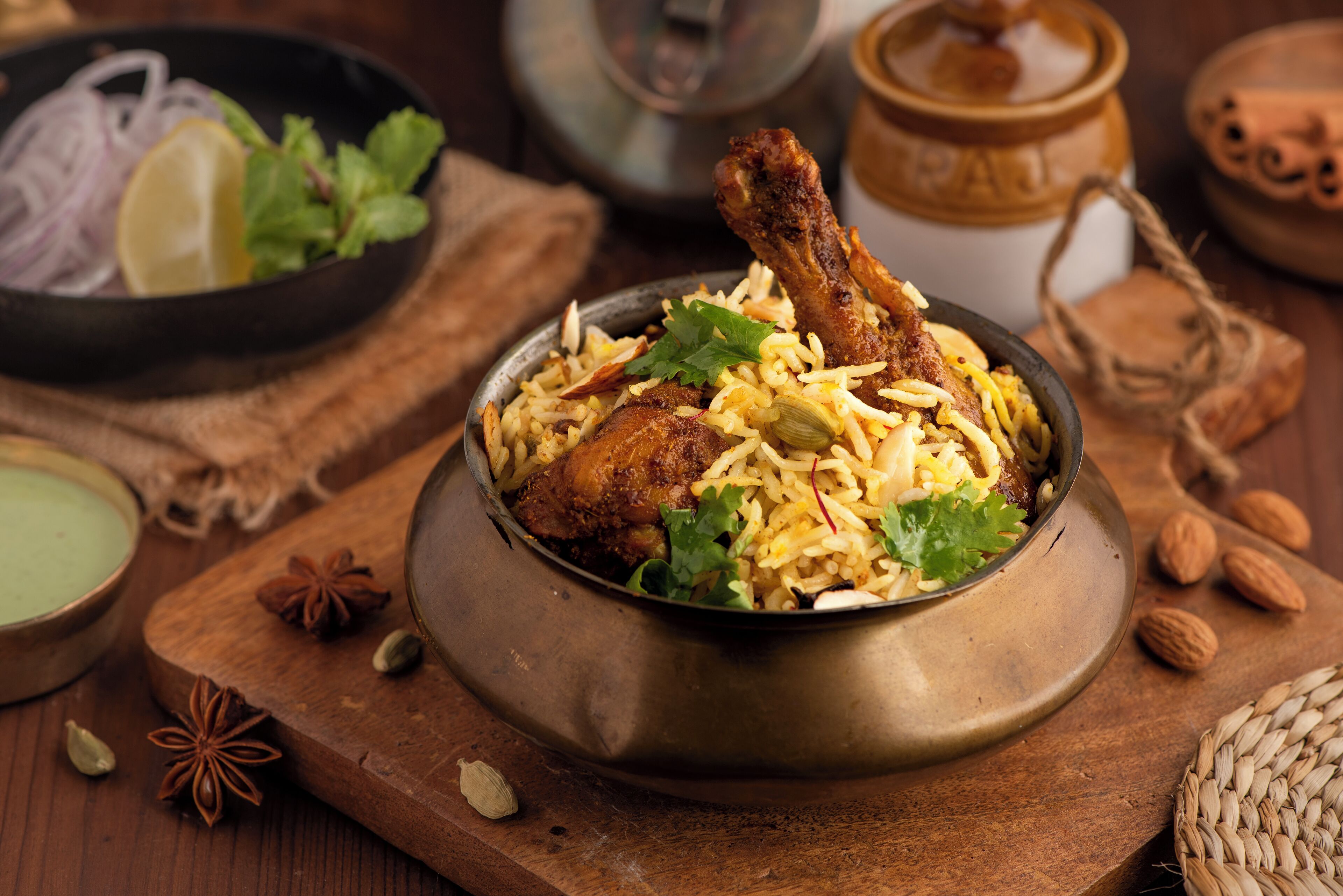Ein traditionelles Biryani-Gericht mit einem großen Fleischstück, garniert mit Kräutern, serviert in einer Metallschüssel auf einer Holzplatte.