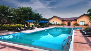 A swimming pool with lounge chairs and umbrellas, surrounded by a building with a tiled roof. - WelcomHeritage Tadoba Vanya Villas Resort & Spa (Chimur)