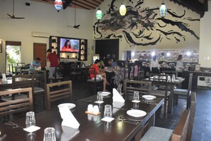 A restaurant with wooden tables and chairs, a TV screen, and a mural on the wall. - WelcomHeritage Tadoba Vanya Villas Resort & Spa (Chimur)