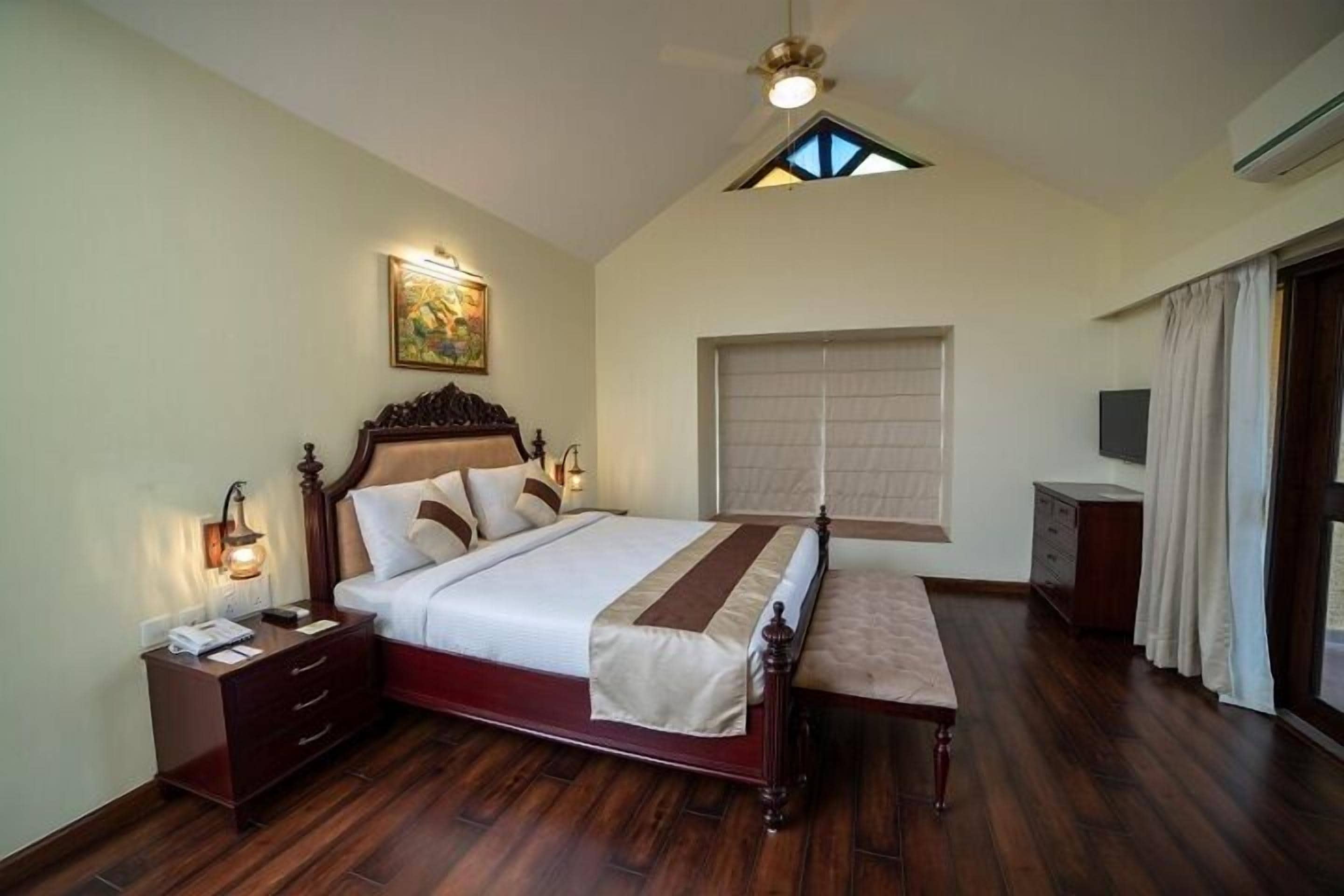Une chambre à coucher avec un grand lit, une tête de lit en bois, une table de chevet, un banc, un ventilateur de plafond, une fenêtre avec des stores et un tableau au mur.
