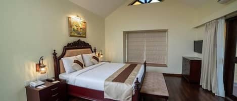 A bedroom with a large bed, a wooden headboard, a nightstand, a bench, a ceiling fan, a window with blinds, and a painting on the wall.