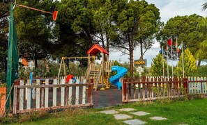 Un parque infantil con un tobogán y columpios, rodeado de árboles y un letrero de hotel.