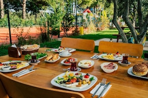 A wooden table set with various dishes, including bread, salads, and a jug of tea, in an outdoor setting with trees and a playground in the background. - Helen Troya Hotel Geyikli (Ezine)