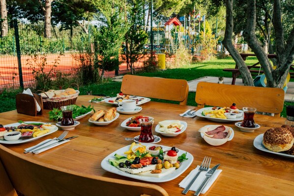 A wooden table set with various dishes, including bread, salads, and a jug of tea, in an outdoor setting with trees and a playground in the background. - Helen Troya Hotel Geyikli (Ezine)
