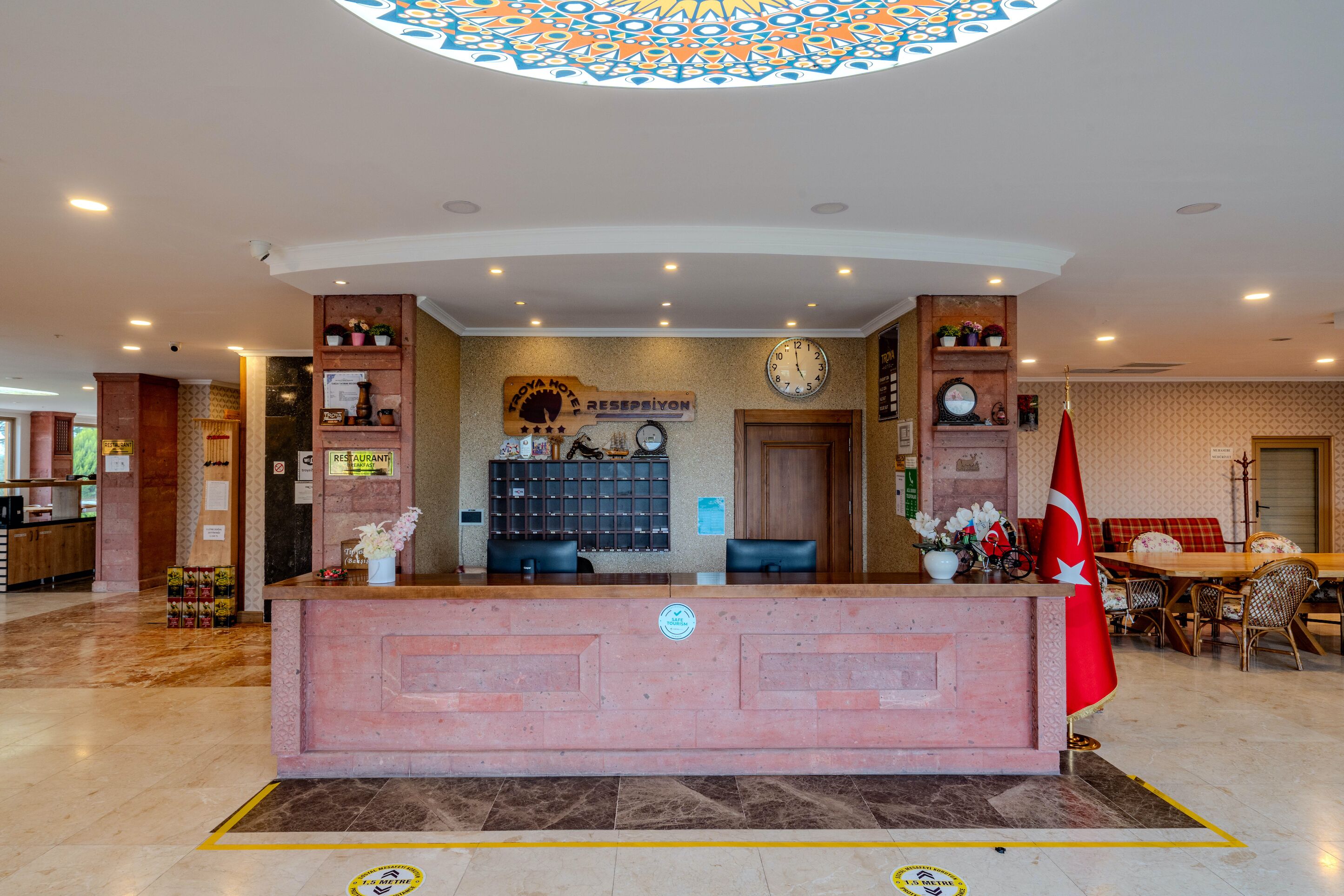 A hotel reception area with a reception desk, a clock, a flag, and a seating area with a table and chairs.