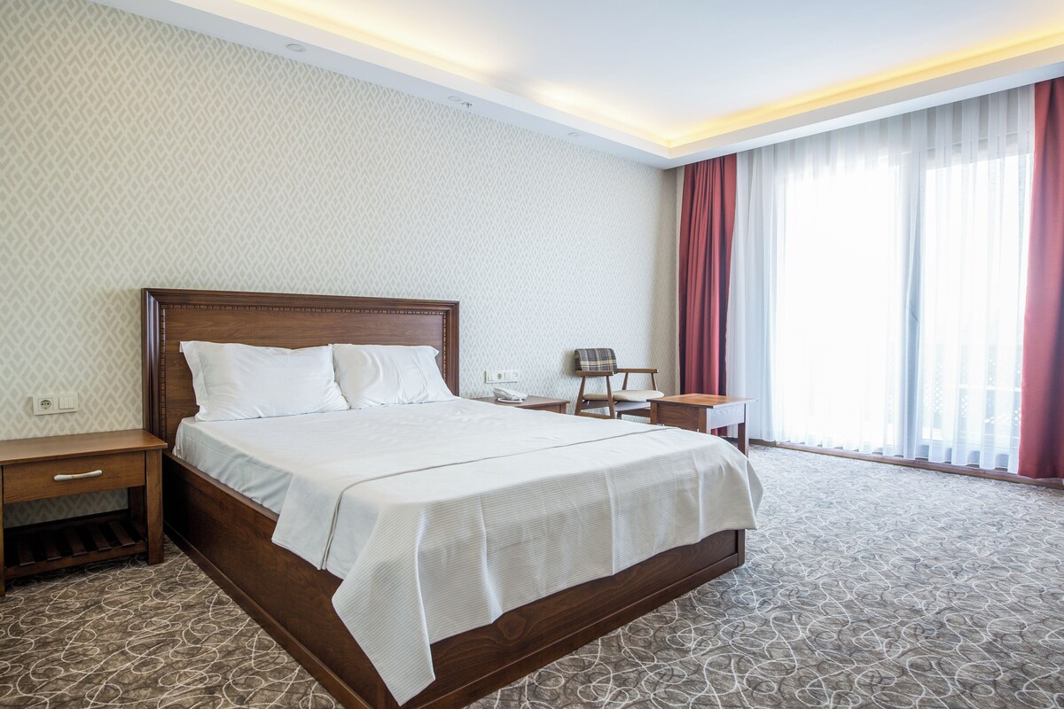 Una habitación de hotel con una cama grande, mesitas de noche, una silla, un escritorio y una ventana con cortinas.