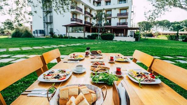 A dining table set outdoors with various dishes, a bowl of bread, and a building in the background. - Helen Troya Hotel Geyikli (Ezine)