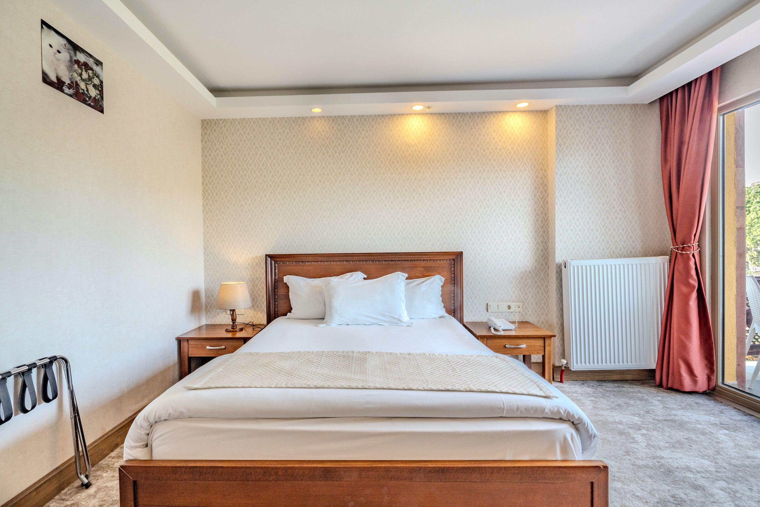 A hotel room with a bed, bedside tables, a lamp, a radiator, and a window with curtains.