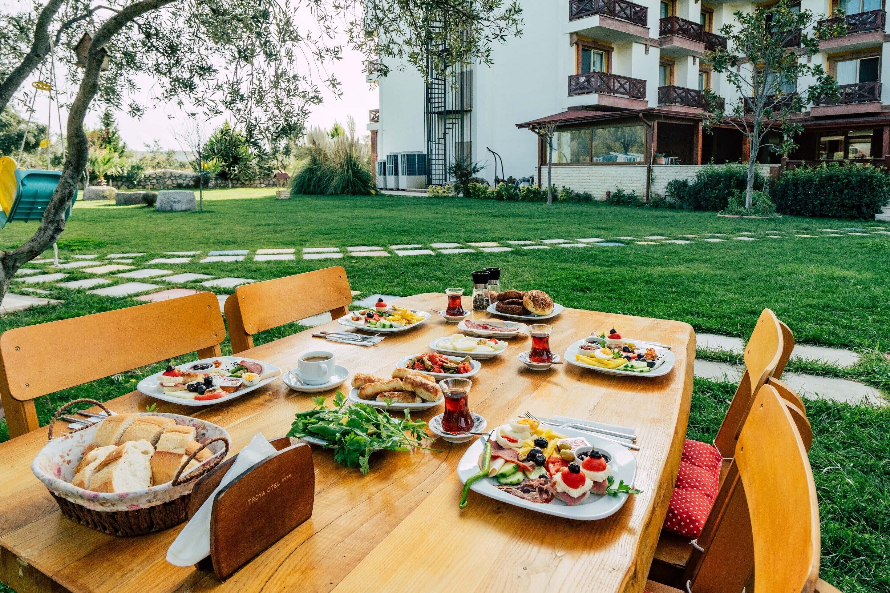 Una mesa de madera con diversos platos y una cesta de pan, rodeada de sillas, en un jardín con un edificio al fondo.