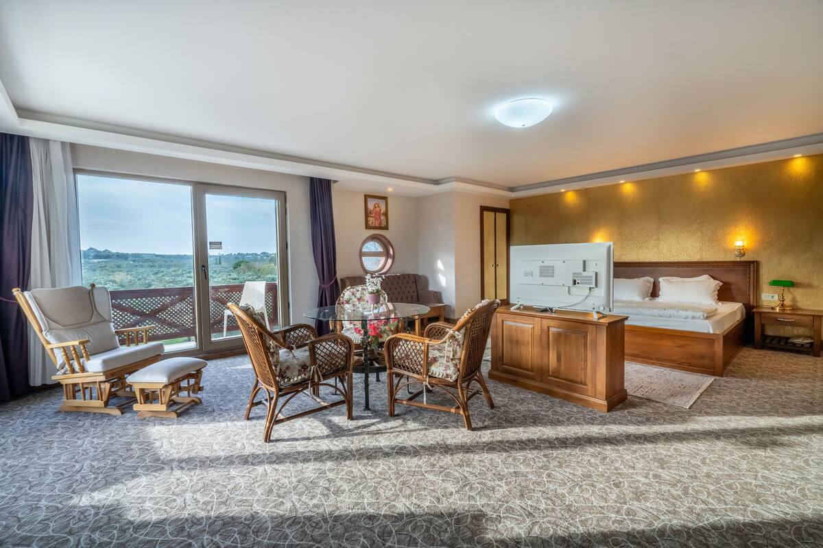 A hotel room with a bed, a desk, a dining table with chairs, a large window with a view, and a balcony.