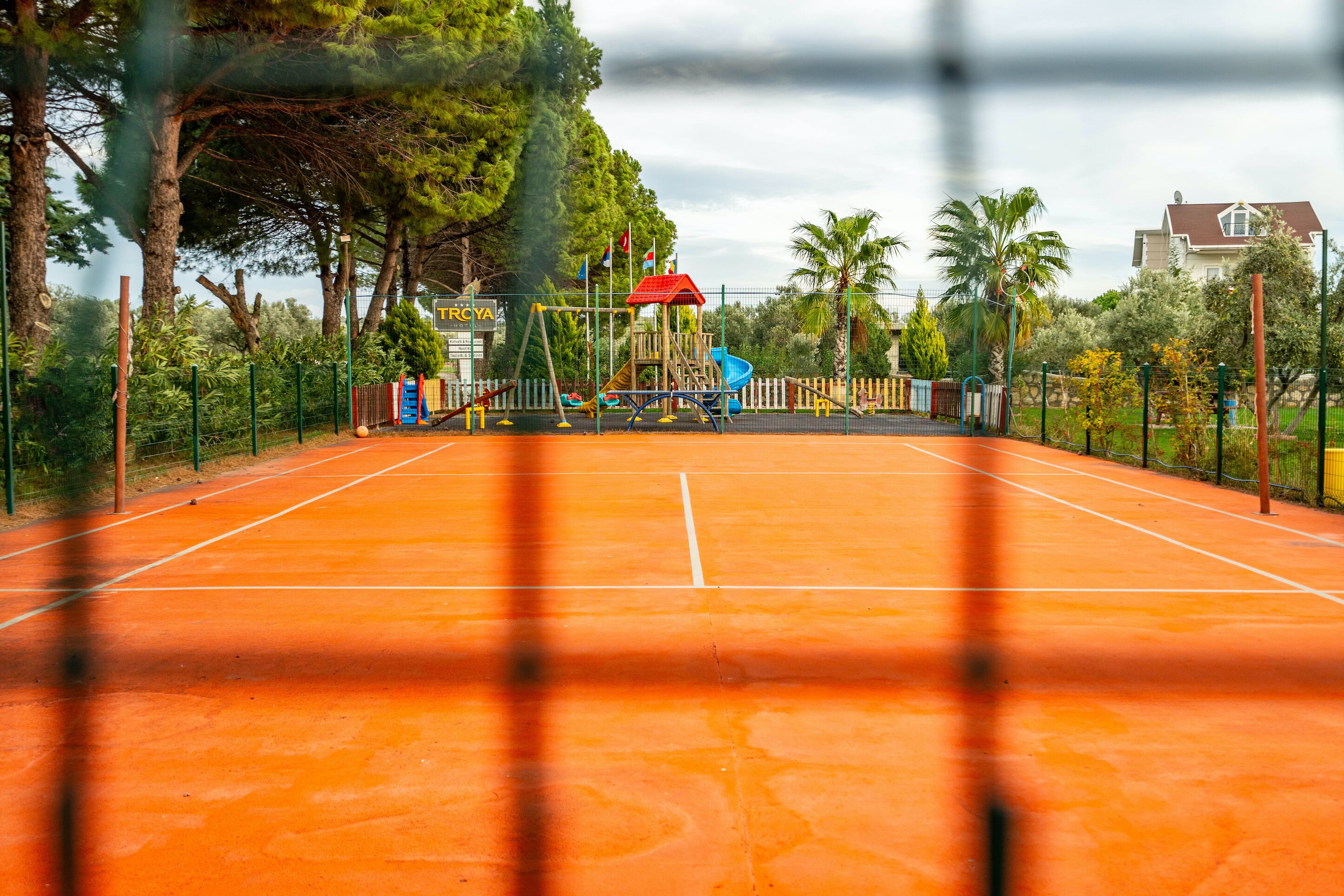 A tennis court with a playground in the background, surrounded by trees and a fence.
