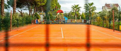 A tennis court with a playground in the background, surrounded by trees and a fence.