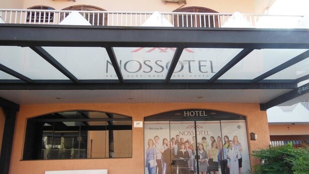 Exterior del hotel Nossotel con una zona de recepción, un banco blanco y un cartel grande con huéspedes.