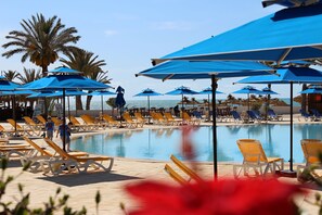 A poolside area with sun loungers and blue umbrellas, palm trees, and a clear view of the ocean.