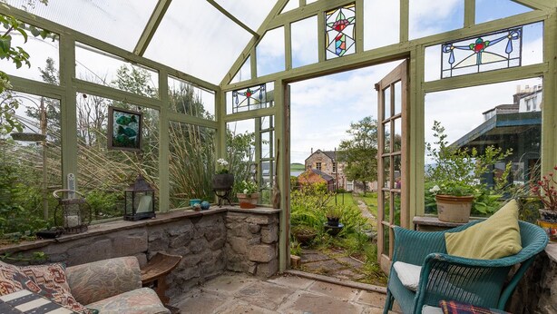 Een oranjerie met een stenen bank, rieten meubels en uitzicht op een tuin en een huis.