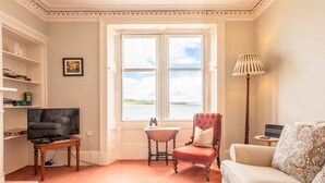 Een woonkamer met een televisie, een rode fauteuil, een witte bank, een lamp en een groot raam met uitzicht op de omgeving.