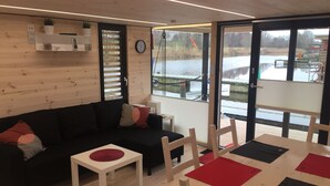 A modern living room with a black sofa, white coffee table, and a dining table with red placemats. Large windows offer views of a lake and dock. - Hausboot Resort Nordseeküste (Wilhelmshaven)