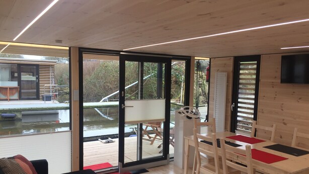 A modern interior with a dining area, a sofa, and a view of a water body outside. - Hausboot Resort Nordseeküste (Wilhelmshaven)