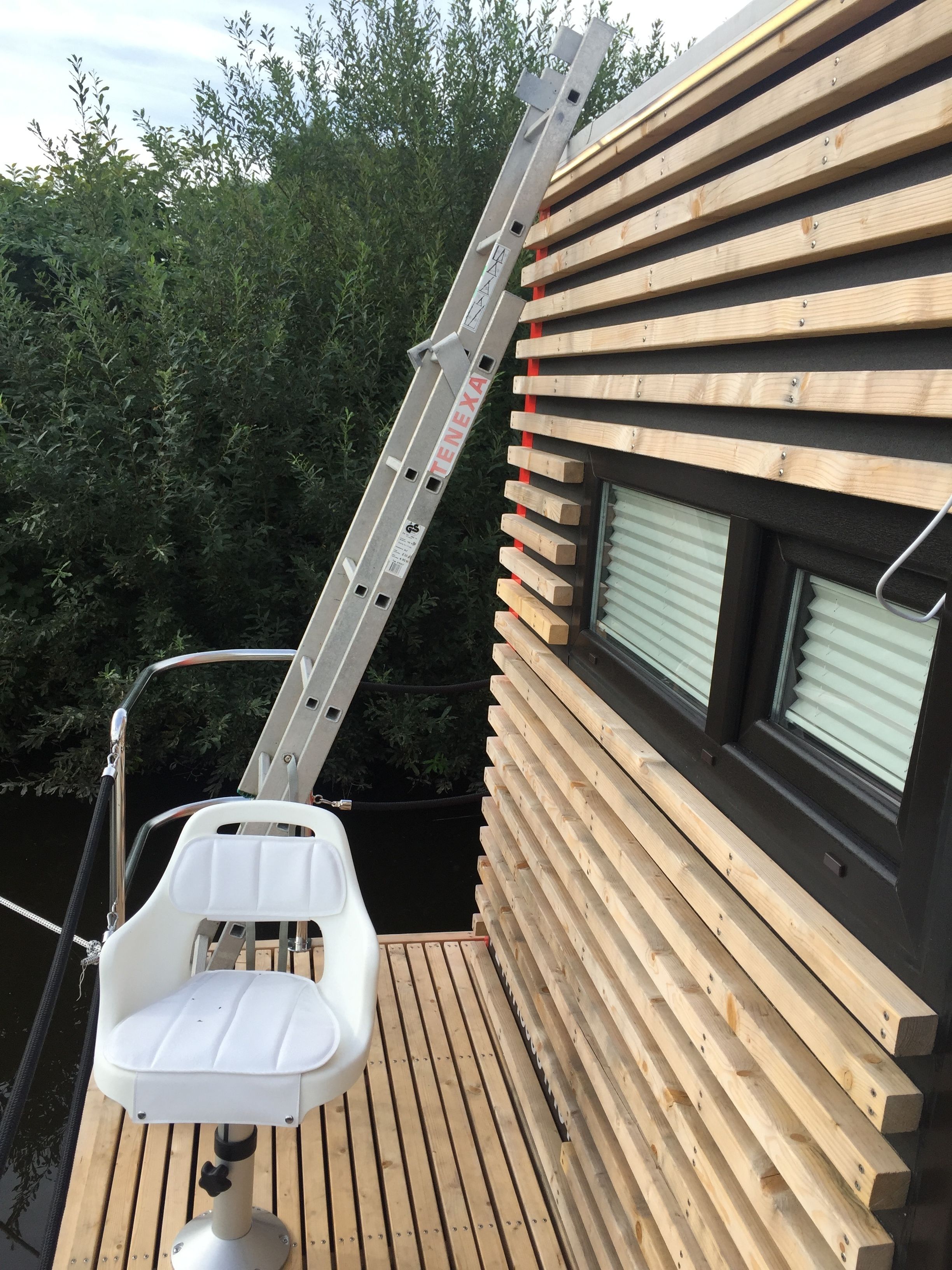 Une terrasse en bois avec une chaise blanche et une échelle en métal appuyée contre un bâtiment dont les murs sont recouverts de planches de bois horizontales et qui possède une fenêtre.