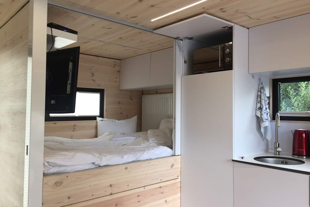 A compact, modern room with a bed, a built-in desk, and a kitchenette.