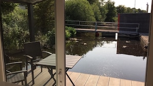 A covered deck with a table and chairs, overlooking a calm water body with a bridge and greenery in the background. - Hausboot Resort Nordseeküste (Wilhelmshaven)