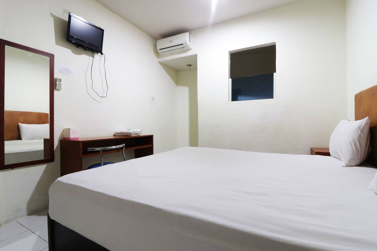 A hotel room with a large bed, a desk with a chair, a television mounted on the wall, an air conditioning unit, and a small window.
