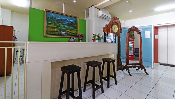 A reception area with a green wall, a painting, a clock, and three stools. - Anno Guesthouse (Makassar)
