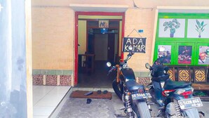 La entrada de un edificio con dos motocicletas estacionadas afuera, un letrero que dice "ADA KAMAR" y una puerta verde decorada con plantas.