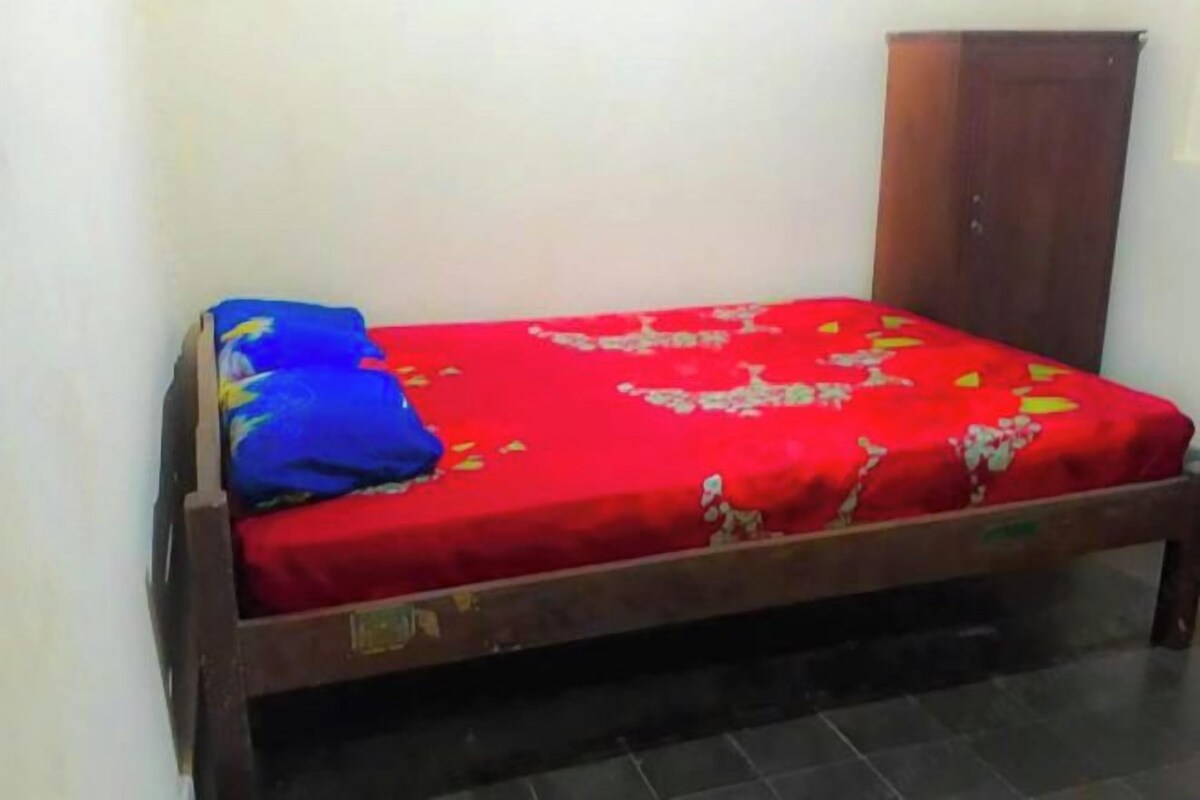 Un llit individual amb un llençol vermell i almoines blaves, un capçal de fusta i un armari al costat d'una paret sense decoració.