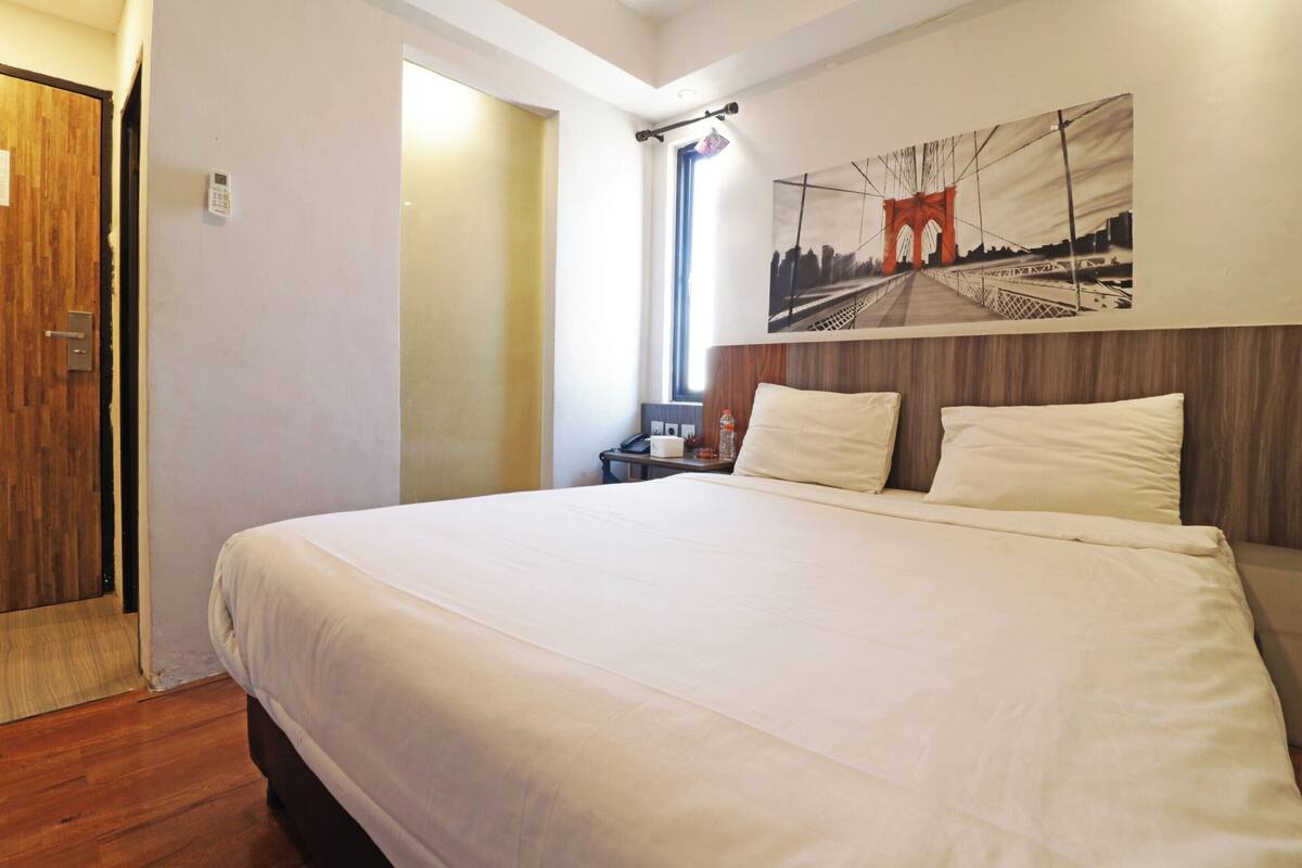A hotel room with a large bed, a wooden door, a small table with a bottle, and a painting of a bridge on the wall.
