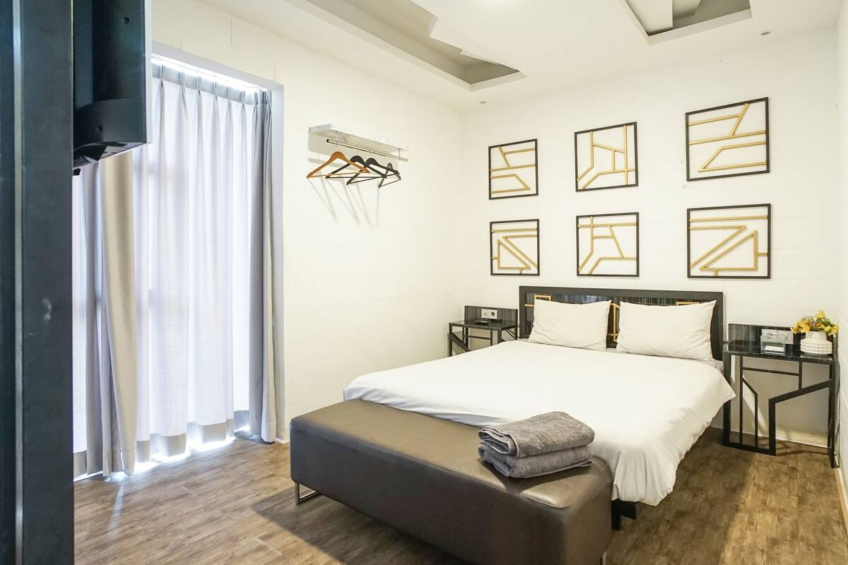 A modern hotel room with a large bed, a bedside table, a nightstand, a bench, a wardrobe, and a window with curtains.