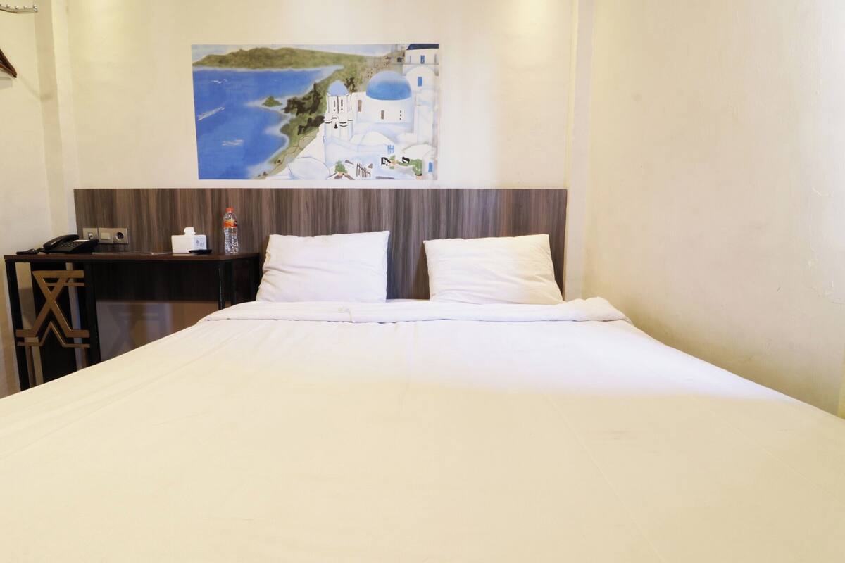 A hotel room with a bed, a nightstand, a painting of a coastal scene, and a wooden headboard.