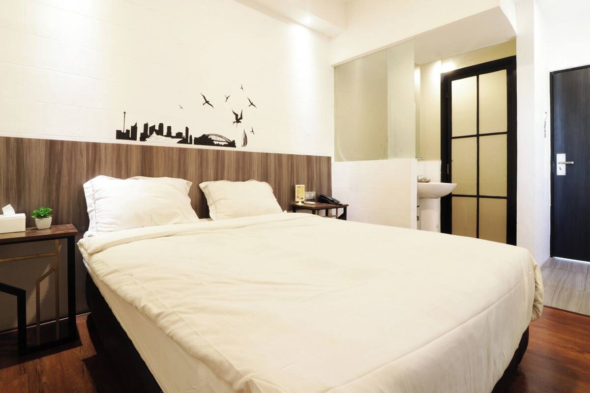 A neatly made bed with a white comforter, a bedside table with a lamp, a wall-mounted cityscape decoration, and a door with a traditional design.