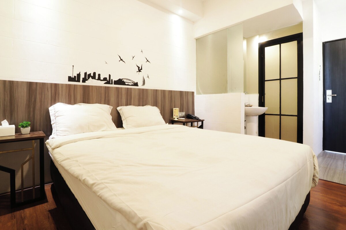 Una cama bien hecha con edredón blanco, una mesita de noche con lámpara, un adorno mural de paisaje urbano y una puerta con diseño tradicional.