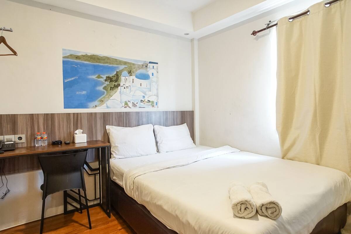 A hotel room with a bed, a desk with a chair, a painting of a coastal scene, and a curtain.