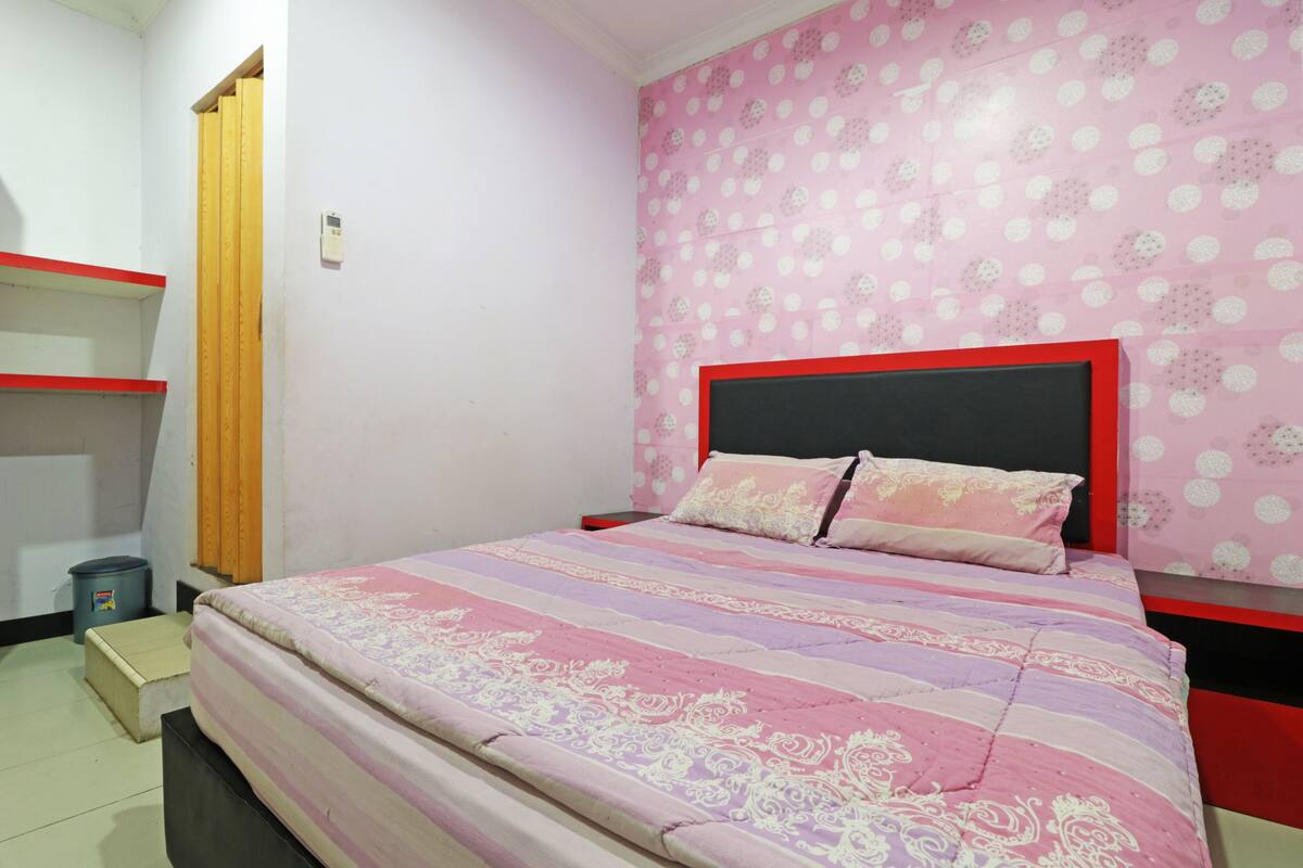Een slaapkamer met een bed, een hoofdeinde met rode lijst, een roze muur met stippen en een bedovertrek met strepen.