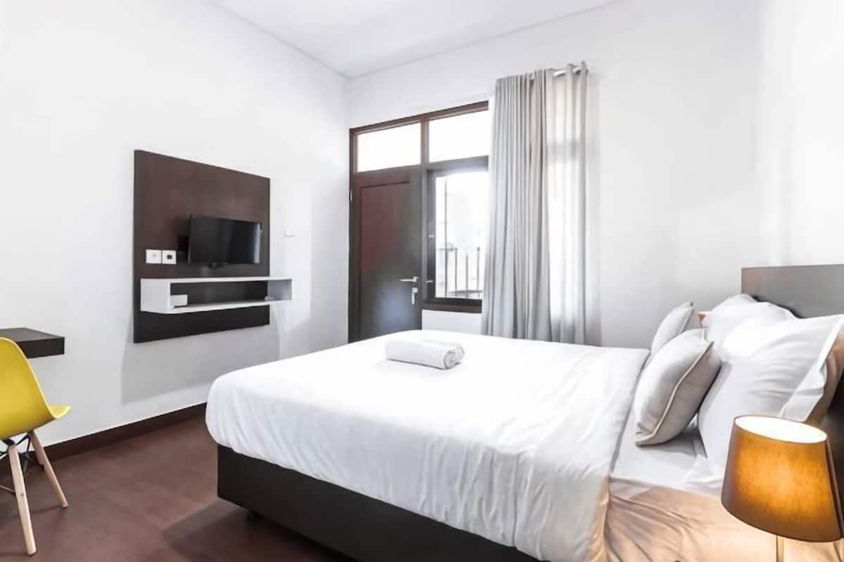 A hotel room with a bed, a TV mounted on the wall, a desk, a chair, and a window with curtains.