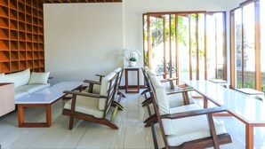 A modern interior with a long dining table, cushioned chairs, a sofa, and a large window offering a view of greenery. - Kak Garden Inn (Kuta)