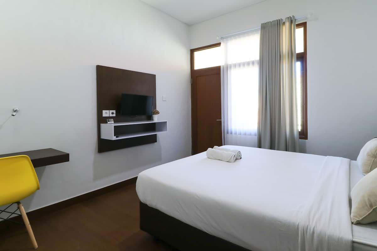 A hotel room with a bed, a TV mounted on the wall, a desk with a chair, and a window with curtains.