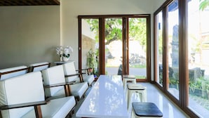 A modern indoor space with a long white table, white cushioned seating, and large windows offering a view of the outdoors. - Kak Garden Inn (Kuta)