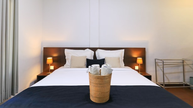 A hotel room with a neatly made bed, two bedside lamps, a bedside table, and a small metal rack. - Kak Garden Inn (Kuta)