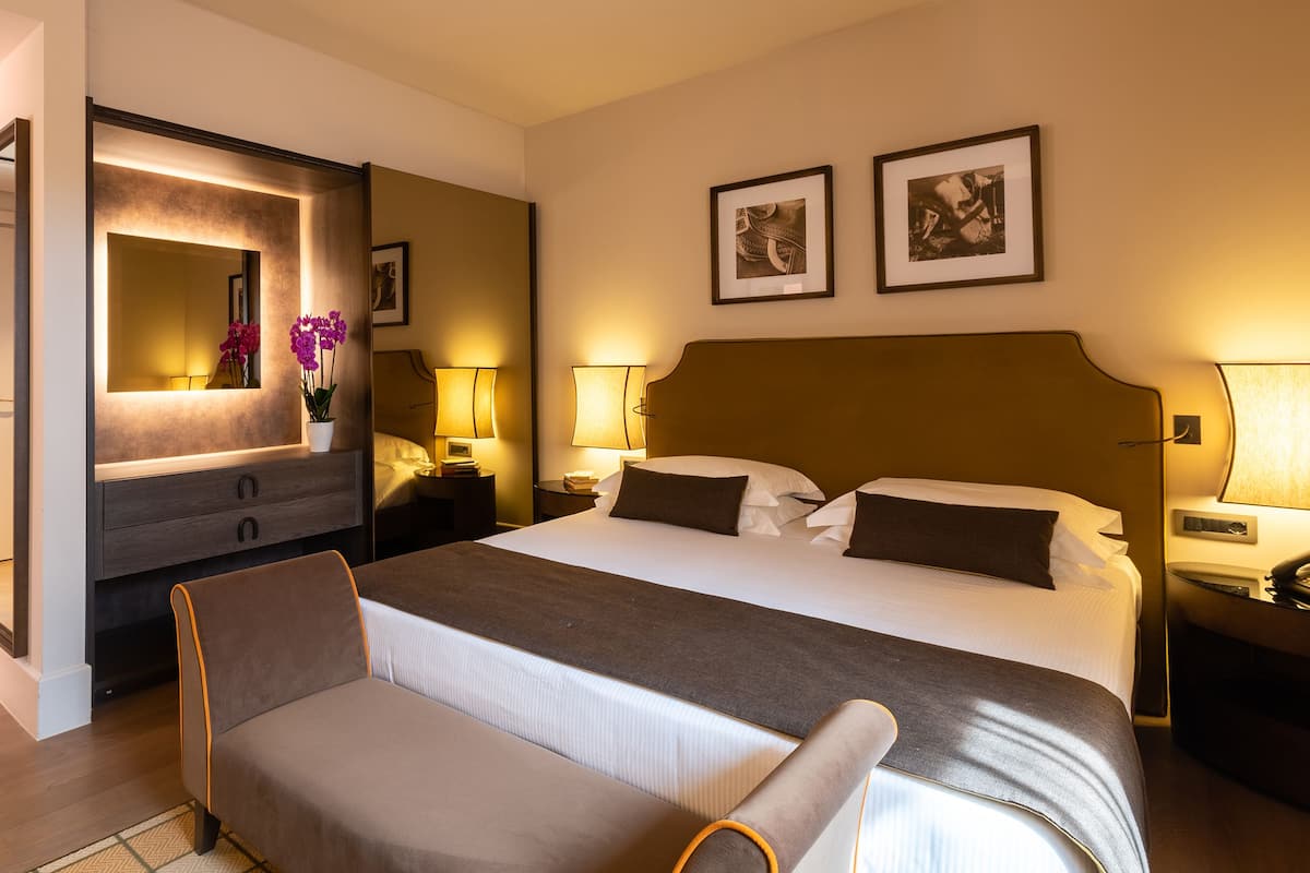 Quarto de hotel com uma cama grande, mesa de cabeceira com abajur, criado-mudo com telefone, uma cadeira, um espelho e duas imagens emolduradas na parede.