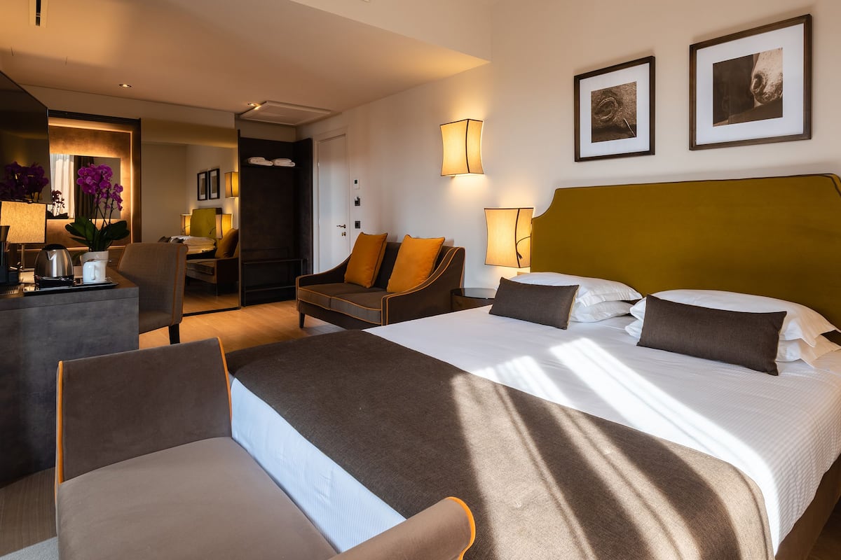 Quarto de hotel com uma cama grande, uma escrivaninha, uma cadeira e um sofá. Há duas quadros na parede.
