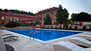 A swimming pool with lounge chairs and umbrellas, surrounded by a brick building with a red roof. - Casa Munfrà (Penango)