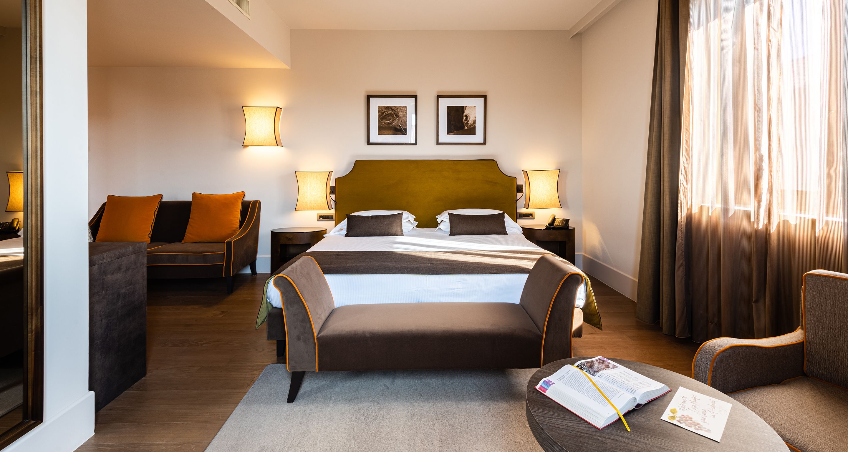 Une chambre d’hôtel avec un grand lit, deux tables de chevet avec des lampes, un canapé et un coin lecture avec un livre et un stylo.