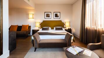 Une chambre d’hôtel avec un grand lit, deux tables de chevet avec des lampes, un canapé et un coin lecture avec un livre et un stylo.