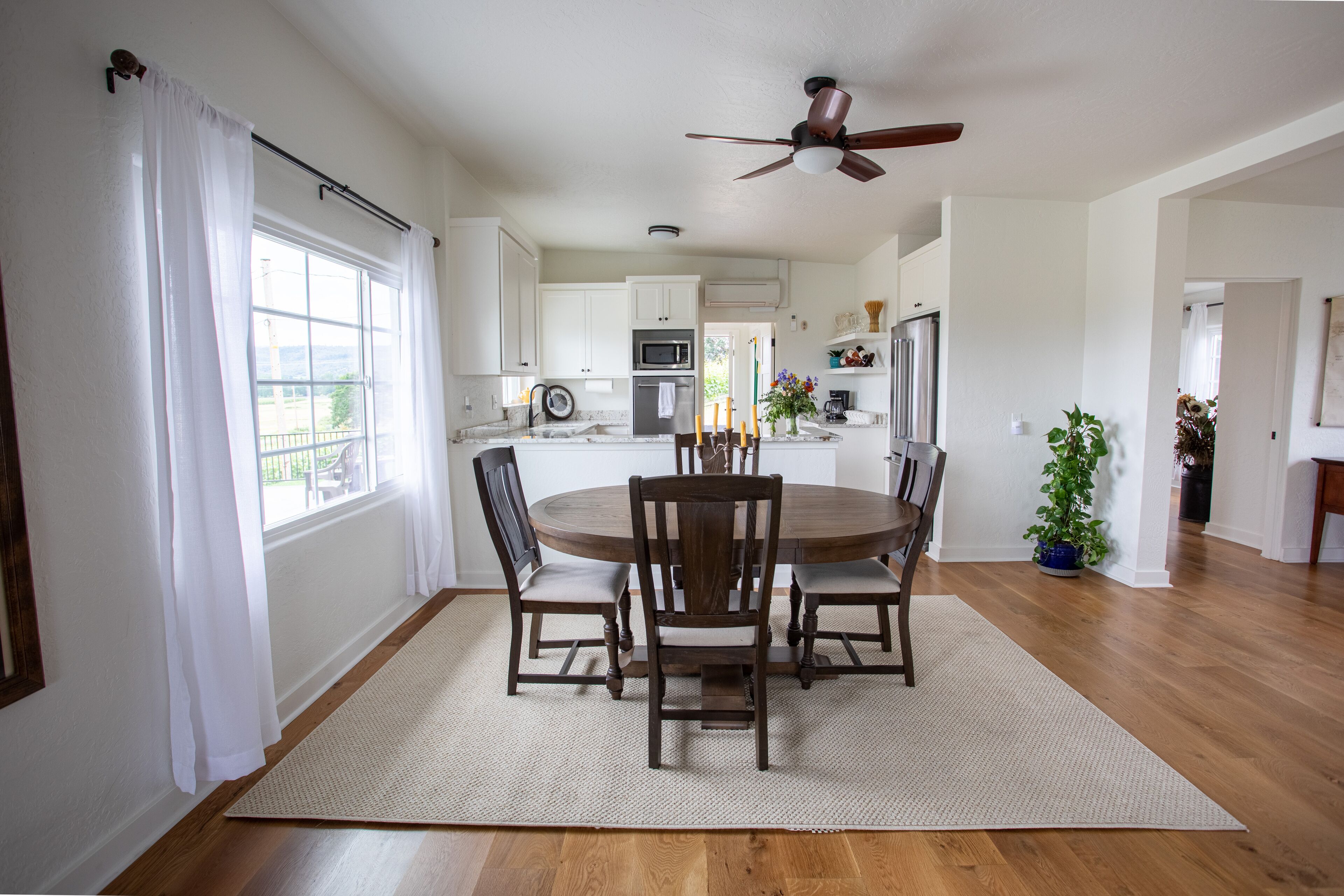 A dining area with a round table, four chairs, a ceiling fan, and a view of the kitchen in the background.